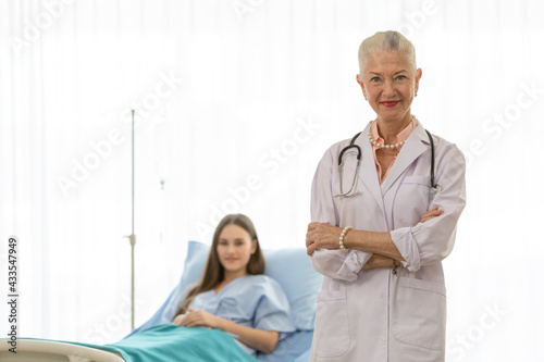 A senior older female doctor standing in an elegant pose  cross her arms  and smile with kindness with a young patient on bed blur in background