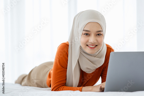 A cute and beautiful Asian Muslim in hijab laying on bed and using tablet notebook computer