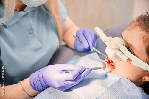 Fotografie Female dentist checking child teeth with dental explorer and mirror while girl lying in chair with inhalation sedation at dental office