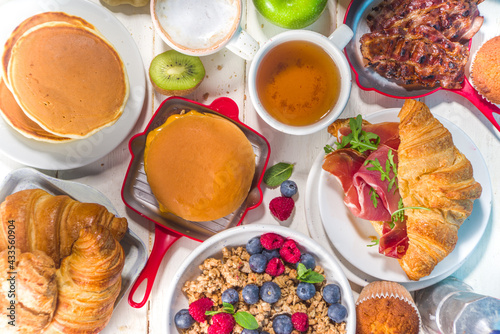 Various traditional breakfast food - fried eggs with bacon, muesli, oats, waffles, pancakes, burger, croissants, fruit berry, coffee, tea and orange juice, white table background copy space top view