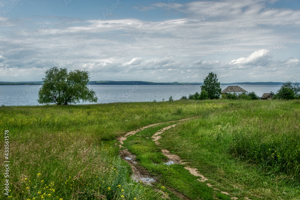 View of the bank of the Kama River (Russia) on a cloudy spring day. A dirt dirt road turns picturesquely to a single wooden house with a vegetable garden. textured white-gray clouds, fresh greens 