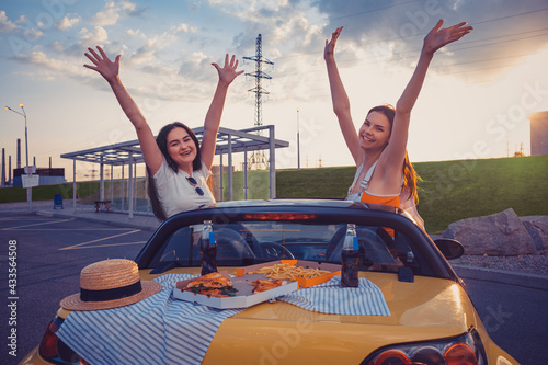 Ladies in casual outfit are smiling, raised up hands, posing in yellow car with french fries, pizza and soda on its trunk. Fast food. Copy space © nazarovsergey