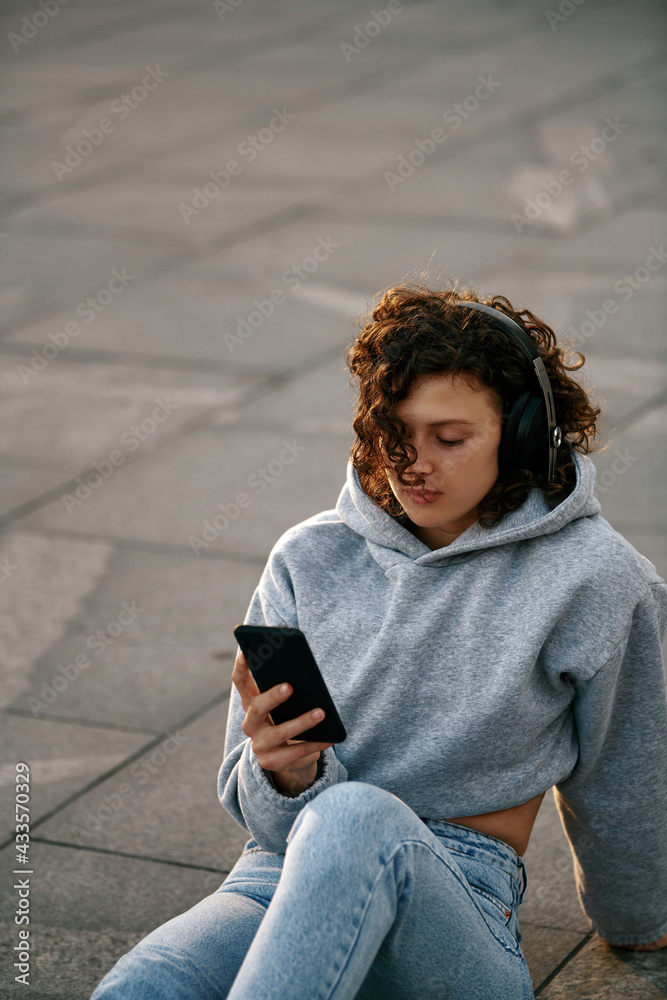 Fototapeta premium Portrait of a young woman with curly hair, dressed casually sitting on the street and listening to music. She is holding the phone in her hands.