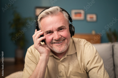 Portrait of a happy man with headphones on his ears. An elderly man laughs into the camera while holding up wireless in-ear headphones. An energetic older man listens to a new playlist of songs.