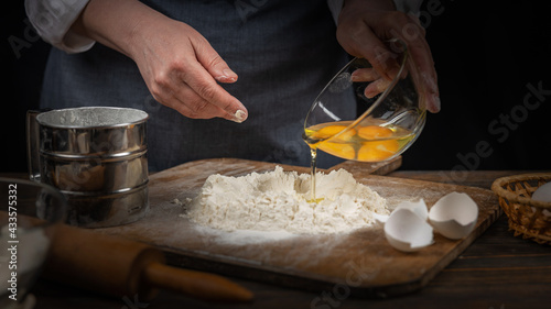 Women's hands, flour and dough. A woman in an apron prepares dough for homemade baking, a rustic home cozy atmosphere, a dark background with unusual lighting.
