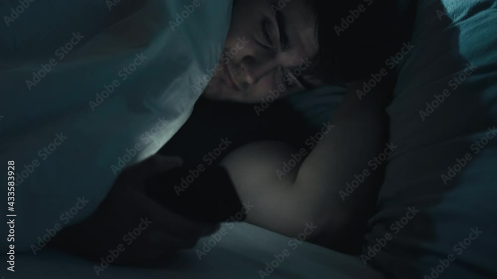 Late online. Night fatigue. Bedtime habit. Home rest. Tired relaxed man with mobile phone falling asleep under blanket in cozy bed in dark bedroom. Late online. Night fatigue. Bedtime habit. Home rest. Tired relaxed man with mobile phone falling asleep under blanket in cozy bed in dark bedroom.