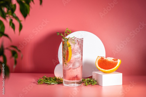 Highball glass of fresh grapefruit cocktail with ice, rosemary and thyme on pink table surface, art drink concept.
