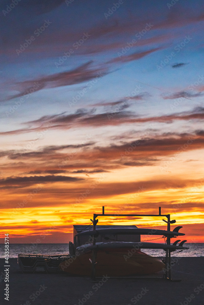 Surf boards silhouetted against vibrant yellow orange sky with clouds at sunset Surf boards silhouetted against vibrant yellow orange sky with clouds at sunset