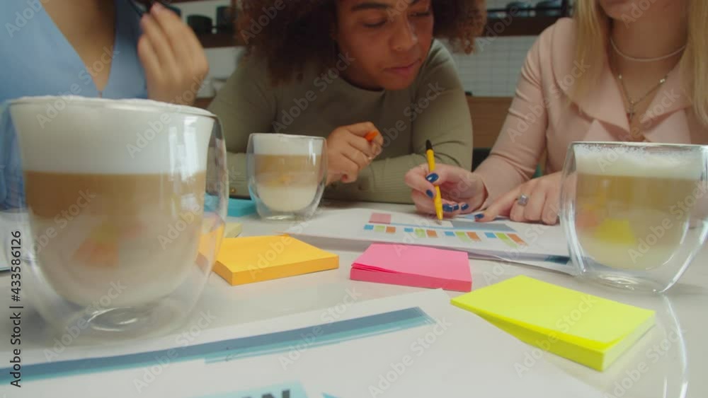 Close-up of successful diverse multiracial female business colleagues analyzing financial data, working on startup project during meeting at cafe with cups of coffee, sticky notes, charts on table. Close-up of successful diverse multiracial female business colleagues analyzing financial data, working on startup project during meeting at cafe with cups of coffee, sticky notes, charts on table.