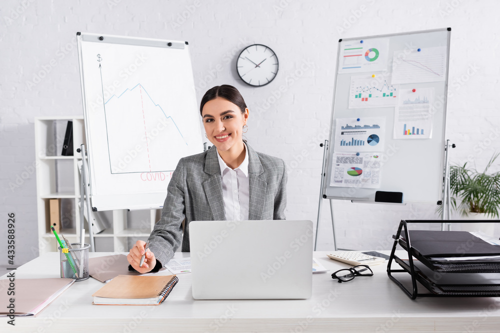 Businesswoman smiling at camera near laptop and papers in office