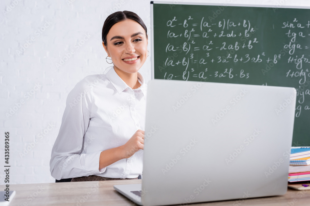 Positive teacher looking at blurred laptop in classroom Positive teacher looking at blurred laptop in classroom