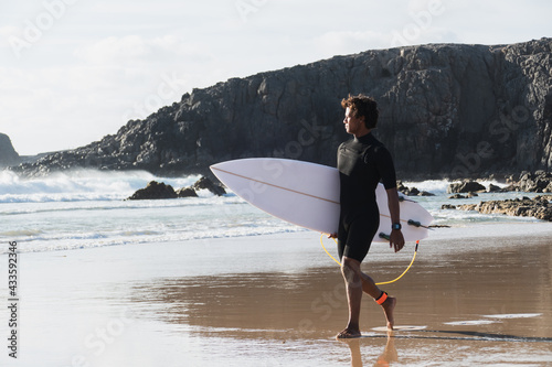 Young surfer walking by the beach shore watching the waves holding surfboard