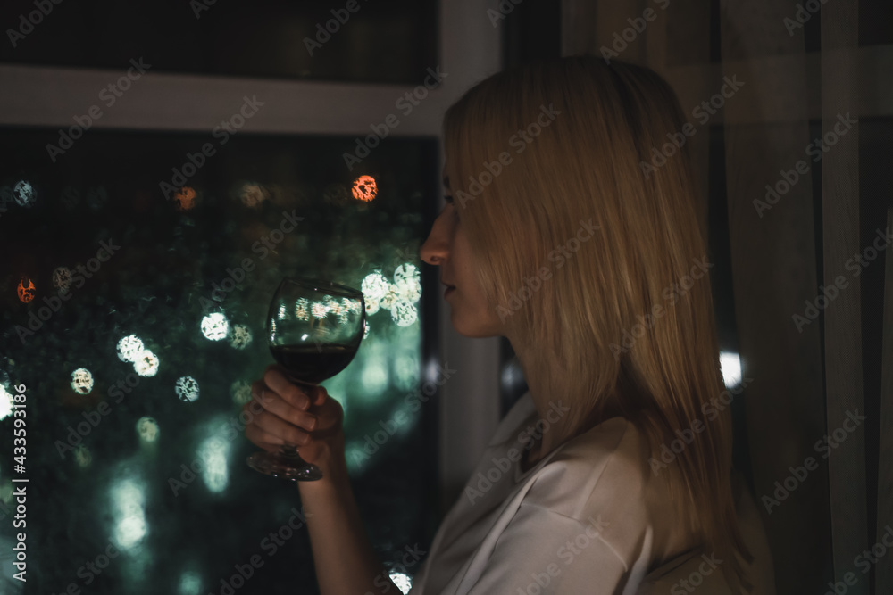 Portrait of a young blonde woman in a white shirt with a glass of red wine looking at the night city through the window. Selective Focus Portrait of a young blonde woman in a white shirt with a glass of red wine looking at the night city through the window. Selective Focus