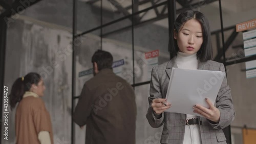 Wallpaper Mural Low angle medium POV of young joyful Asian businesswoman standing in busy modern office, holding pile of documents, then looking up and smiling on camera Torontodigital.ca