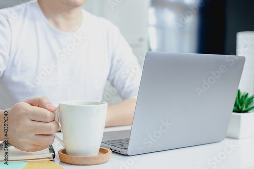 Man in a white shirt picks up a coffee mug and thinks of work on his laptop while working at home.