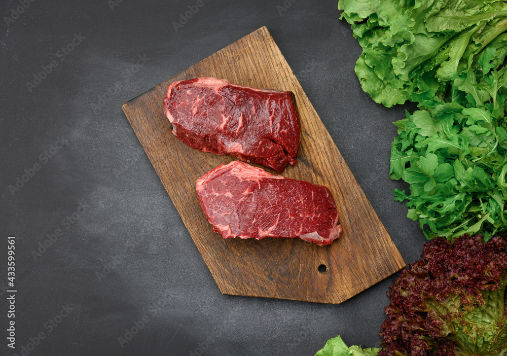 two raw pieces of beef steak on a brown wooden board, black table, next to green lettuce leaves