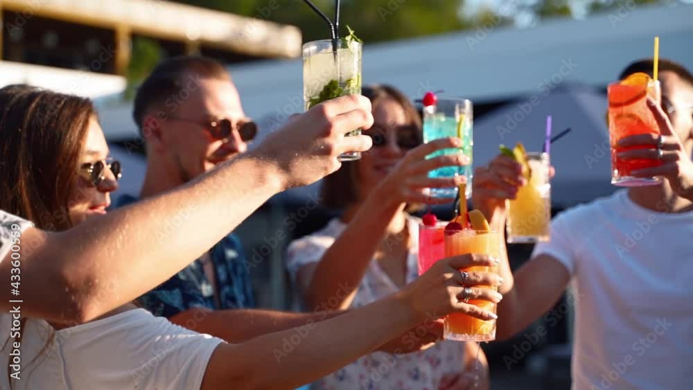 Close view of friends having fun at poolside summer party, clinking glasses with fresh summer cocktails near hotel swimming pool outdoors. People toast drinking juice at luxury villa in slow motion.