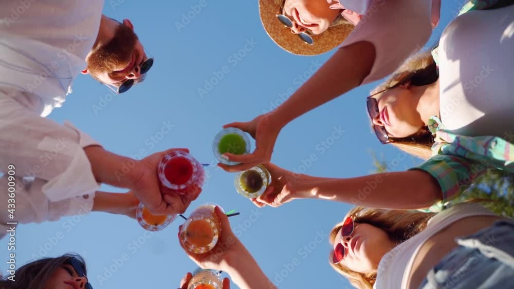 Low angle view of friends having fun at pool party, clinking glasses with colorful summer cocktails near hotel swimming pool. People toast drinking fresh juice at luxury summer villa in slow motion.