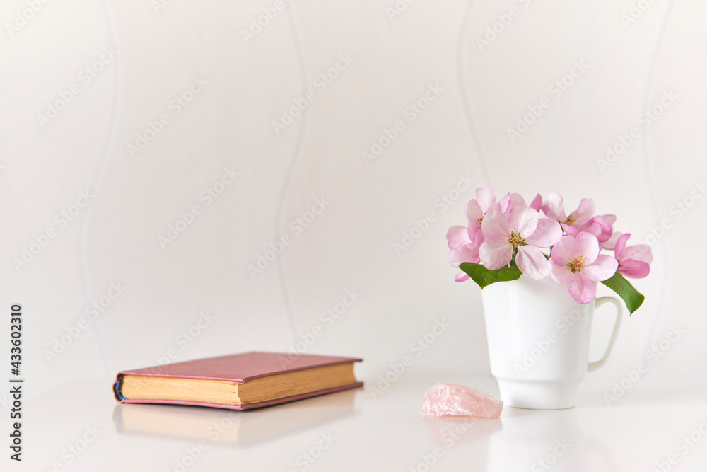 Gentle bouquet of pink apple tree flowers in a white coffee cup, pink mineral, small poetry book on a white coffee table Gentle bouquet of pink apple tree flowers in a white coffee cup, pink mineral, small poetry book on a white coffee table