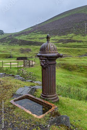 Old rusty cast iron drinking fountain with decorative Lion Head pattern and a ceramic basin sunk into ground at Wanlockhead in the Leadhills