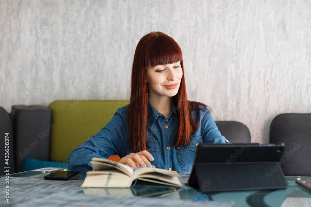 Portrait of young pleasant Caucasian red haired woman, in jeans shirt, sitting at the table in cafe or coworking, reading the book and typing on tablet pc. Study, freelance work, communication Portrait of young pleasant Caucasian red haired woman, in jeans shirt, sitting at the table in cafe or coworking, reading the book and typing on tablet pc. Study, freelance work, communication