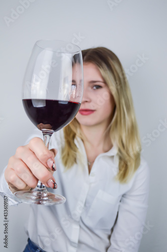 Portrait of a beautiful young blonde woman who raises a glass of red wine to appreciate its color and nuances at a wine tasting - Focus on the glass of wine