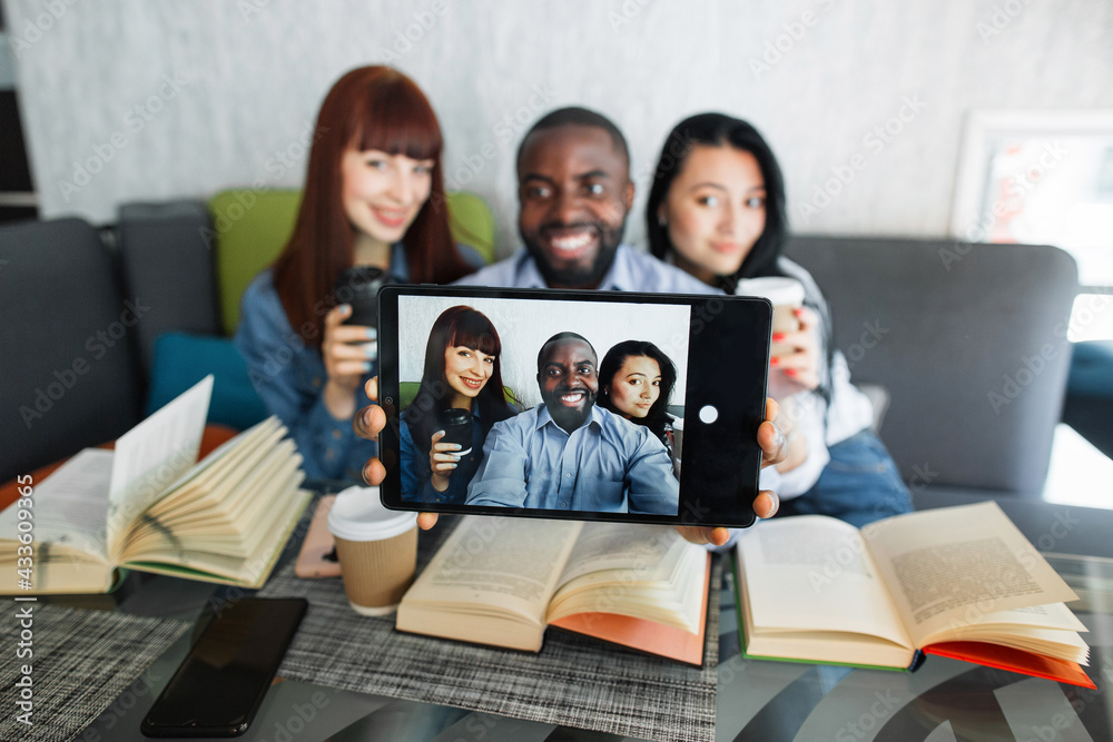Fototapeta premium Young cheerful multiracial friends taking selfie in cafe or college campus, using digital tablet. Multiethnic students, sitting at the table with books and making self portrait on tablet pc