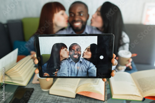 Three multiethnic friends, enjoying time for reading books in cafe together, taking selfie photo on dogotal tablet. African man grinning to camera while Caucasian and Asian girl kissing him on cheeks photo