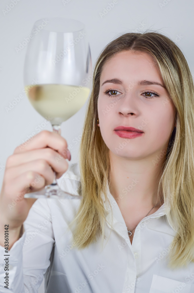 Portrait of a beautiful young blonde woman who raises a glass of white wine to appreciate its color and nuances at a wine tasting - Focus on her face Portrait of a beautiful young blonde woman who raises a glass of white wine to appreciate its color and nuances at a wine tasting - Focus on her face
