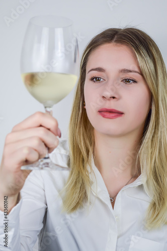 Portrait of a beautiful young blonde woman who raises a glass of white wine to appreciate its color and nuances at a wine tasting - Focus on her face