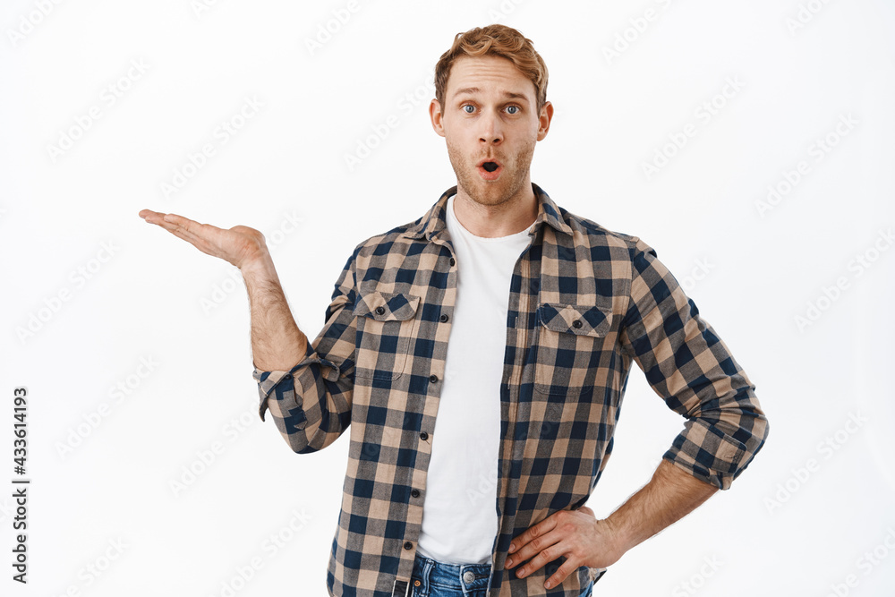 Impressed redhead man say wow while holding an item, raising hand with opened palm, showing an object, product in his arms, standing over white background Impressed redhead man say wow while holding an item, raising hand with opened palm, showing an object, product in his arms, standing over white background
