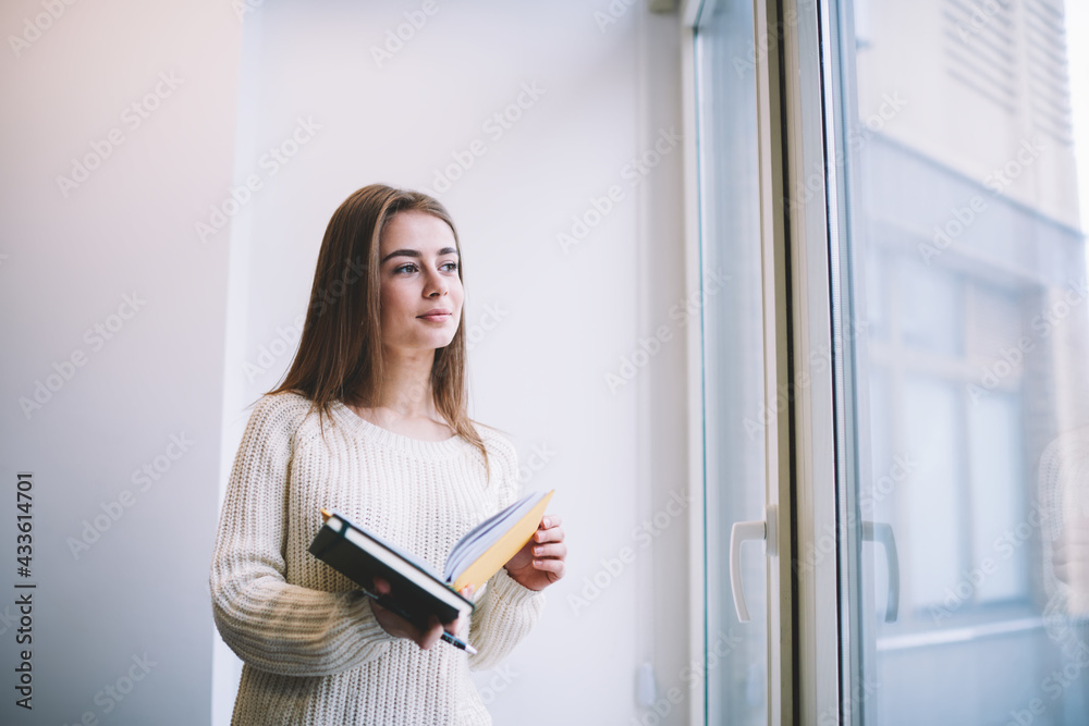 Dreamy young lady standing with copybook and book near window Dreamy young lady standing with copybook and book near window
