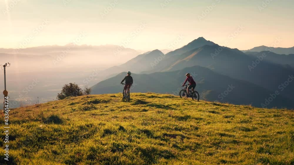 Aerial shot of two female mountain bikers riding across the meadow, stopping at the top of the hill, giving high five
