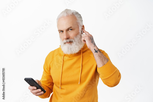 Stylish hipster senior man put wireless earphone in ear, holding smartphone and looking at camera, going to make phone call, listening music in headphones, wearing hoodie, white background