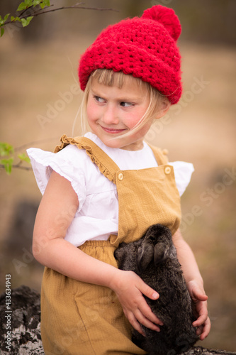 Little girl in a red hat in a spring forest plays with a rabbit in nature