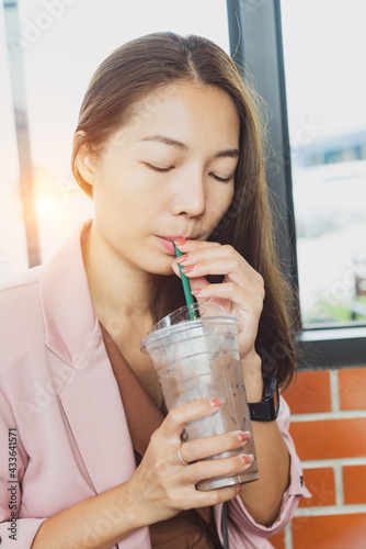 Young attractive businesswoman, successful working in modern pink suit holding glass of cold protein chocolate shake, drink with straw. Smiling Happy woman drinking smoothie cocoa juice in cafe