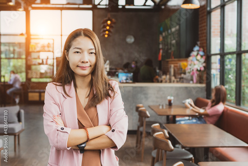 Young Asian businesswoman in pink suit standing with arms folded in coffee shop, confident herself, pride owner of a business modern restaurant. Smiling beautiful cute girl with happy face in cafe