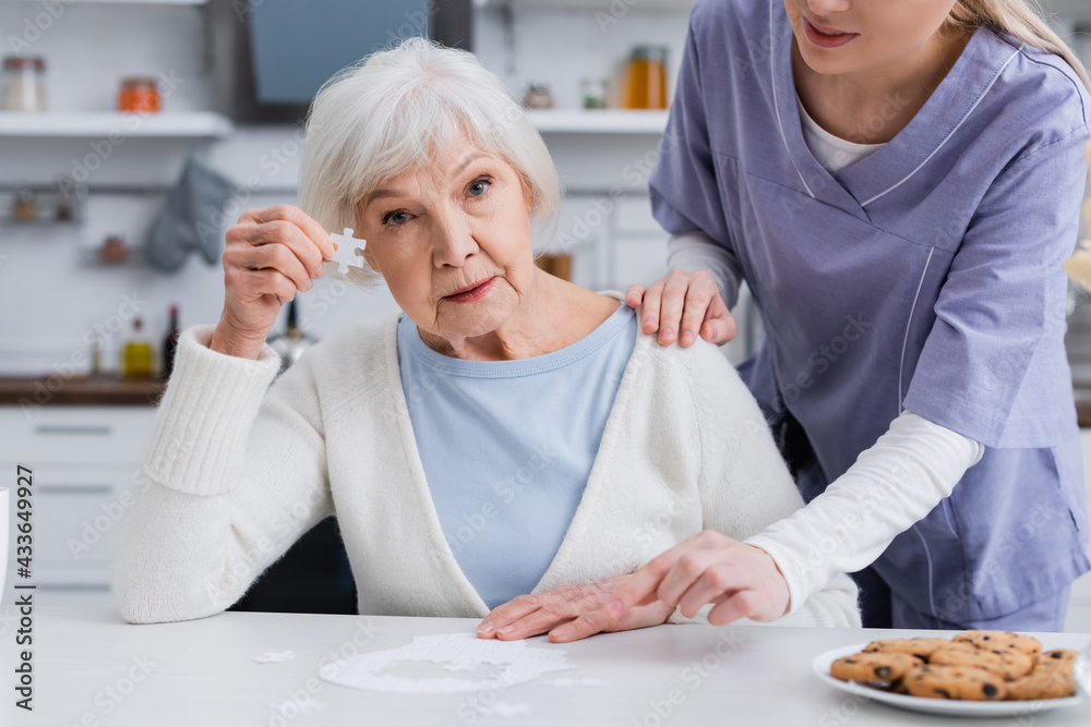 elderly woman showing piece of jigsaw puzzle while nurse touching her shoulder elderly woman showing piece of jigsaw puzzle while nurse touching her shoulder