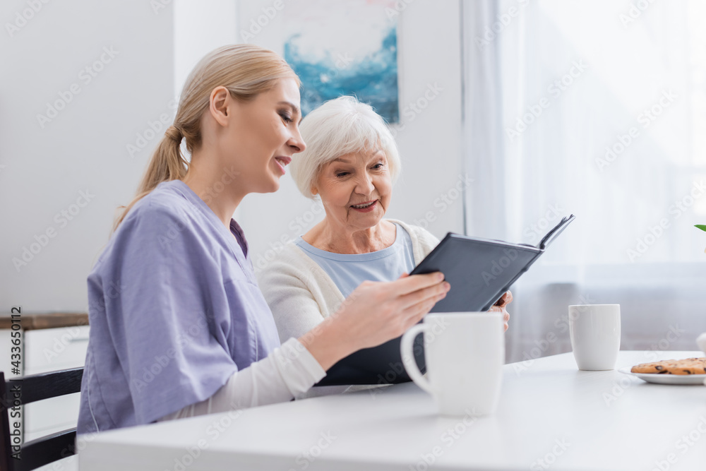 smiling nurse with happy elderly woman looking at photo album near cups of tea smiling nurse with happy elderly woman looking at photo album near cups of tea
