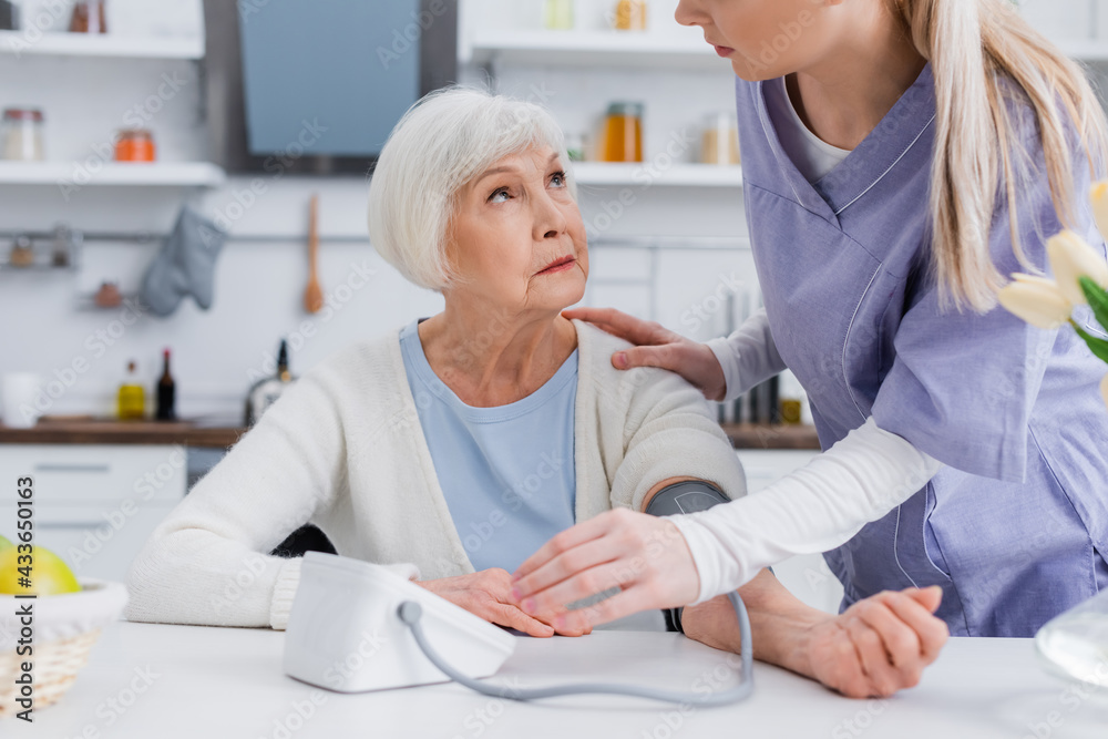 Fototapeta premium social worker touching shoulder of elderly woman while measuring her blood pressure in kitchen