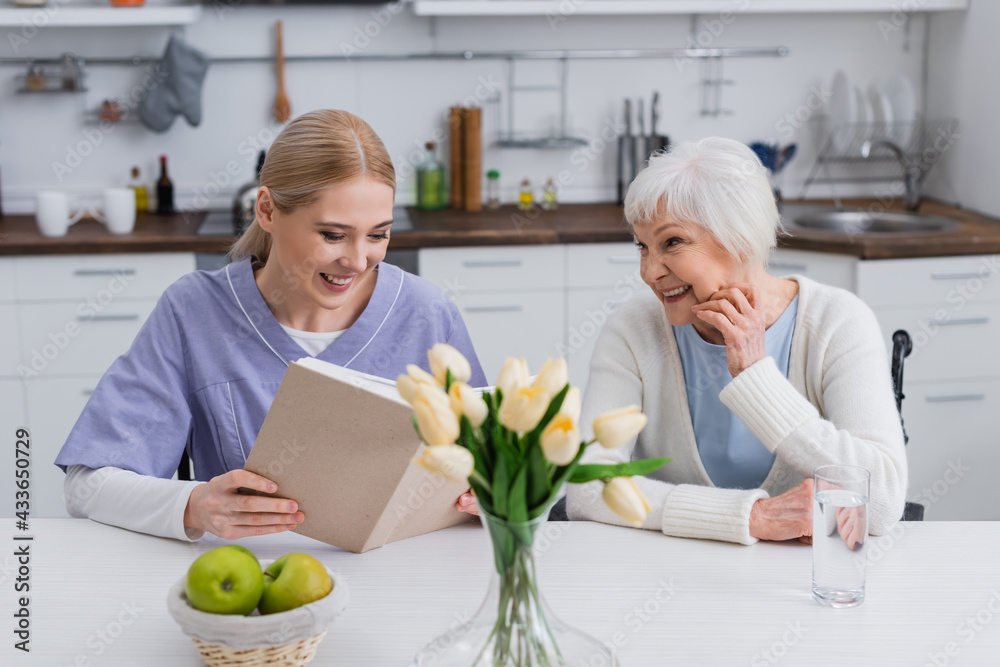 smiling nurse reading book to happy senior woman near fresh apples and tulips in kitchen