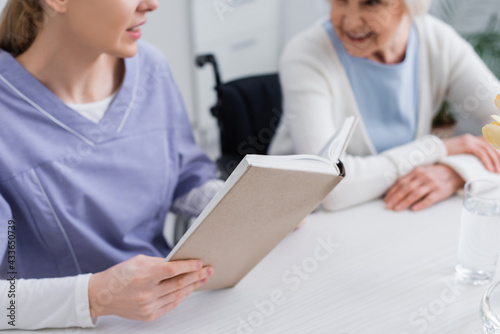 cropped view of young nurse reading book to elderly woman, blurred background