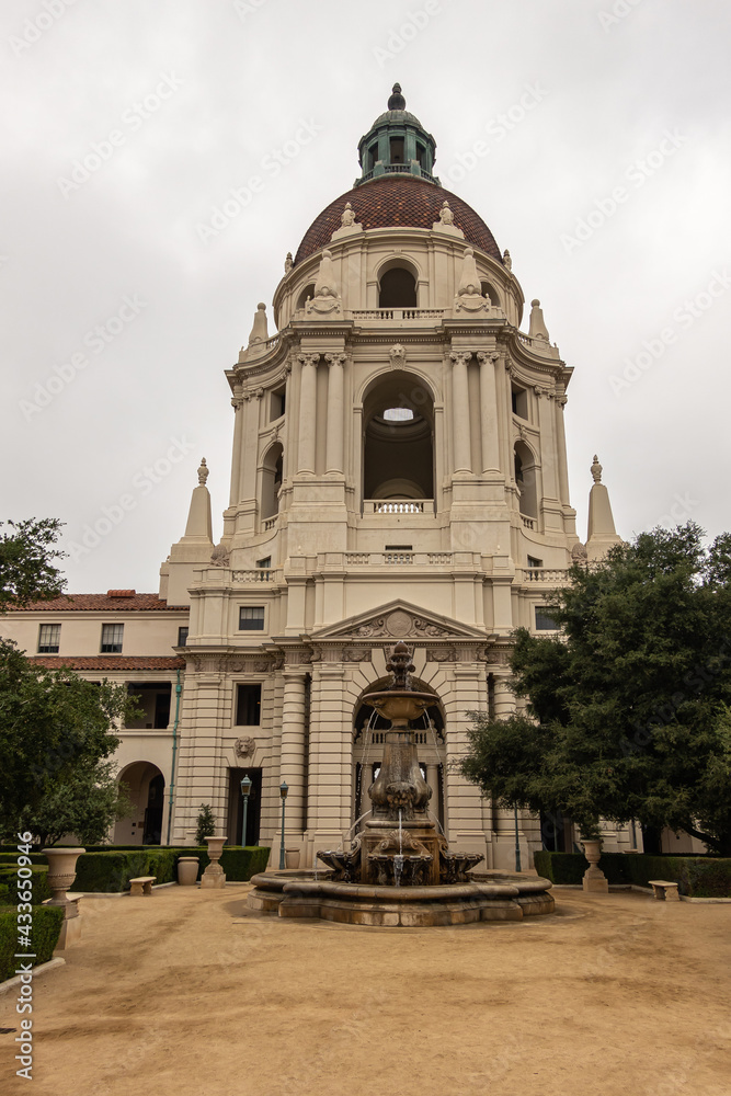 Obraz premium Pasadena, CA, USA - May 11, 2021: Beige stone main tower with dome and fountain in front in sandy courtyard and green foliage garden of historic City Hall under silver sky. 
