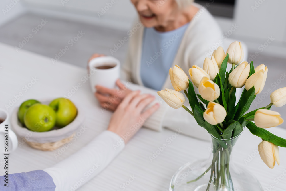 selective focus of fresh tulips near social worker touching hand of elderly woman on blurred background, cropped view