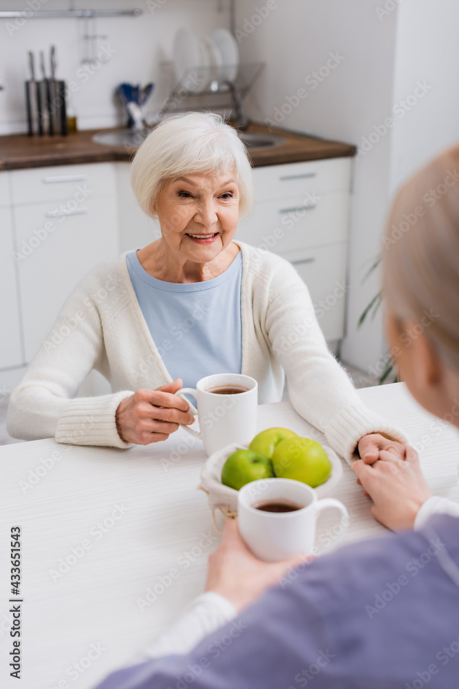 happy elderly woman holding hands with social worker near cups of tea in kitchen, blurred foreground