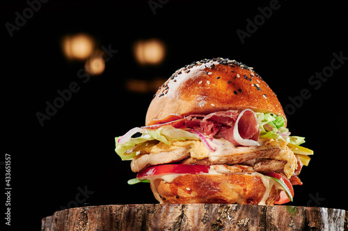 Juicy burger with chicken and bacon, cheese, tomatoes, lettuce on a wooden cut on a black background. Burger menu. Space for text. High quality photo