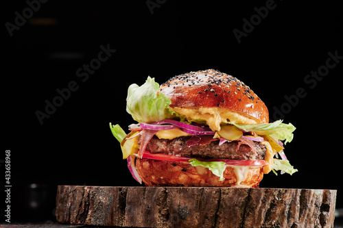 Beef burger with cheese, tomatoes, red onions, cucumber and lettuce on wooden cut on black background. Space for text. Unhealthy food. High quality photo