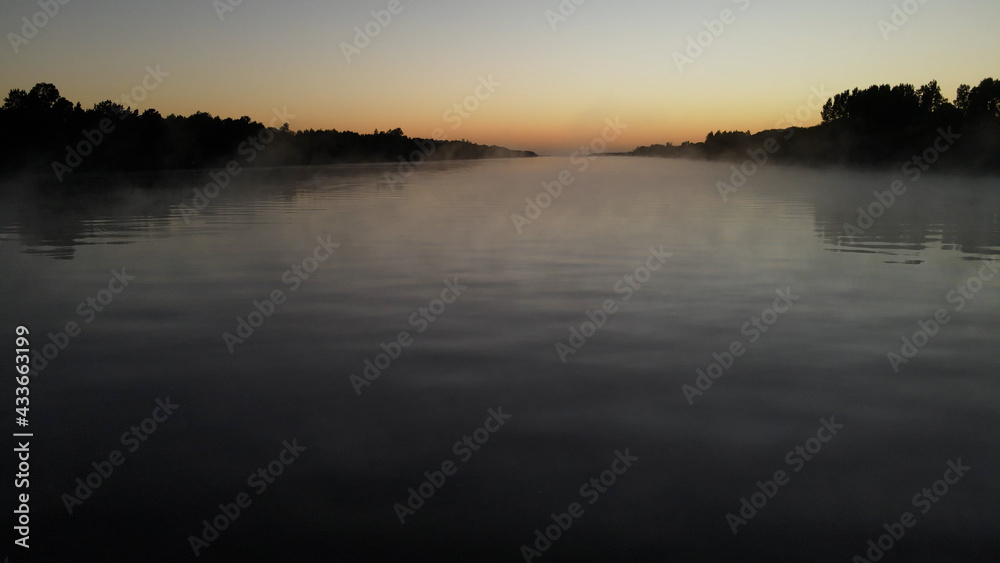 Foggy tranquil amazon river during colorful sunset in background. Silhouette of trees and plants on shore.