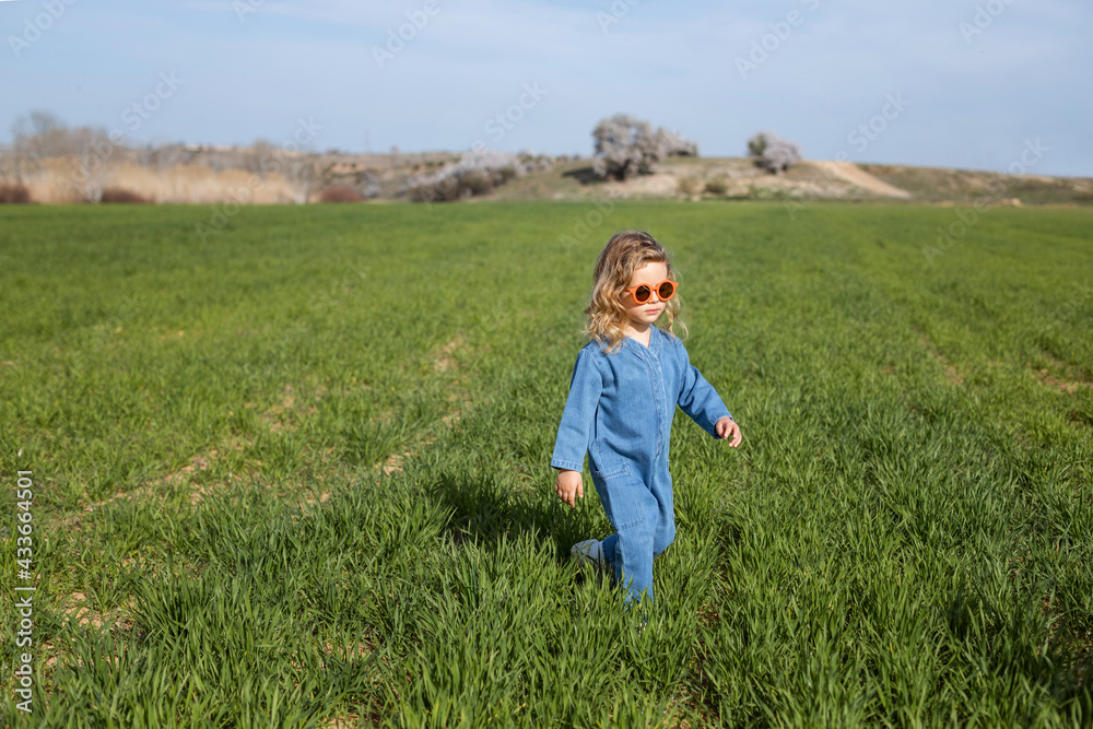 Cute happy little girl in trendy clothes and sunglasses standing and relaxing on grassy lawn Cute happy little girl in trendy clothes and sunglasses standing and relaxing on grassy lawn
