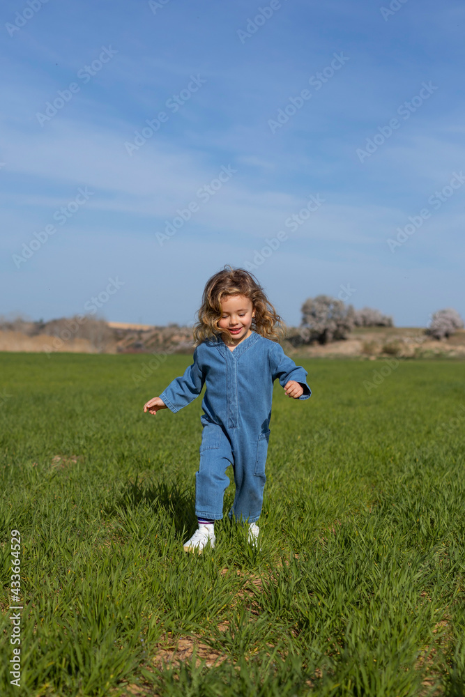 Delighted girl with curly hair smiling and running on green grass against blue sky on summer day in field Delighted girl with curly hair smiling and running on green grass against blue sky on summer day in field